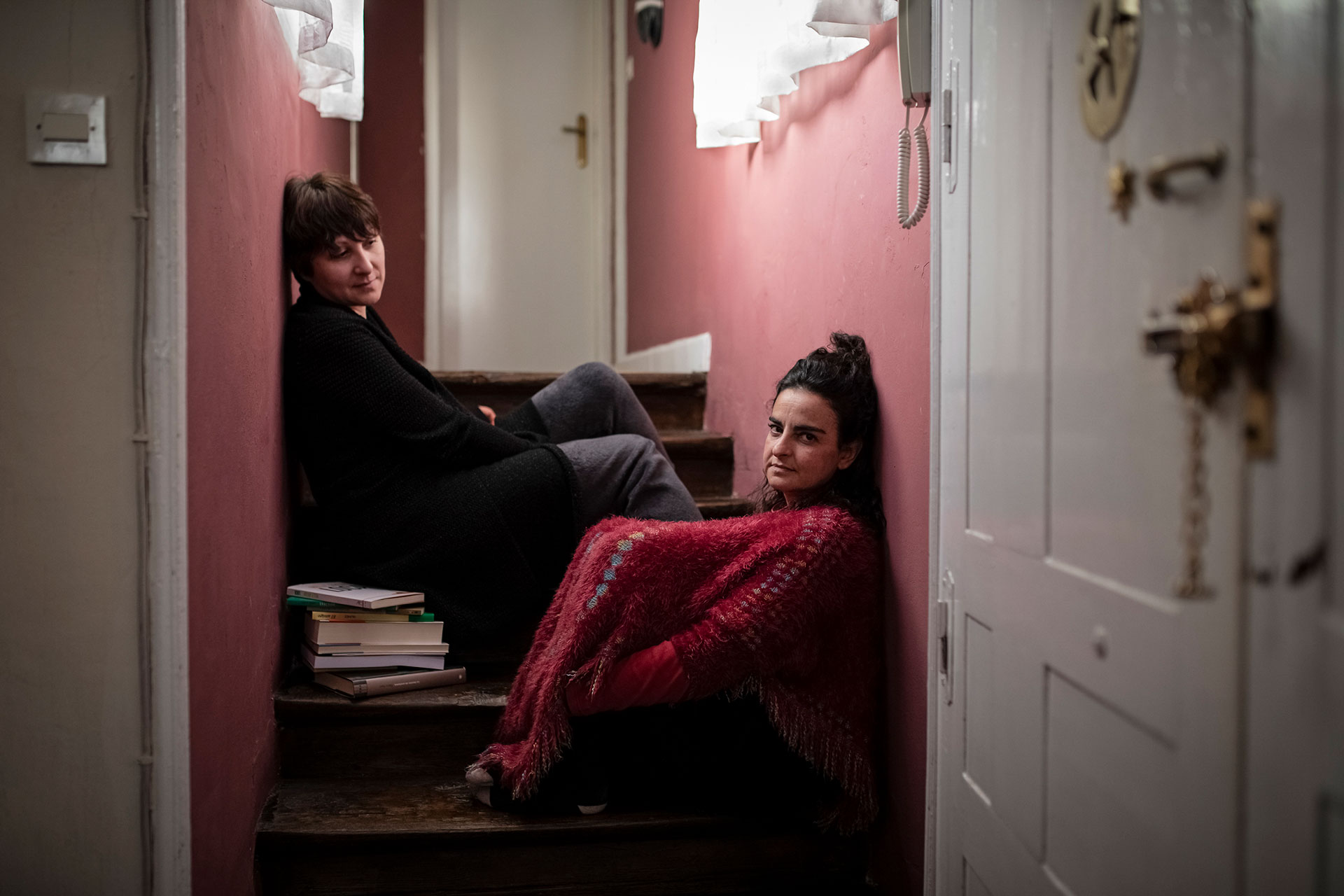 Two women sit on the stairs in their flat.