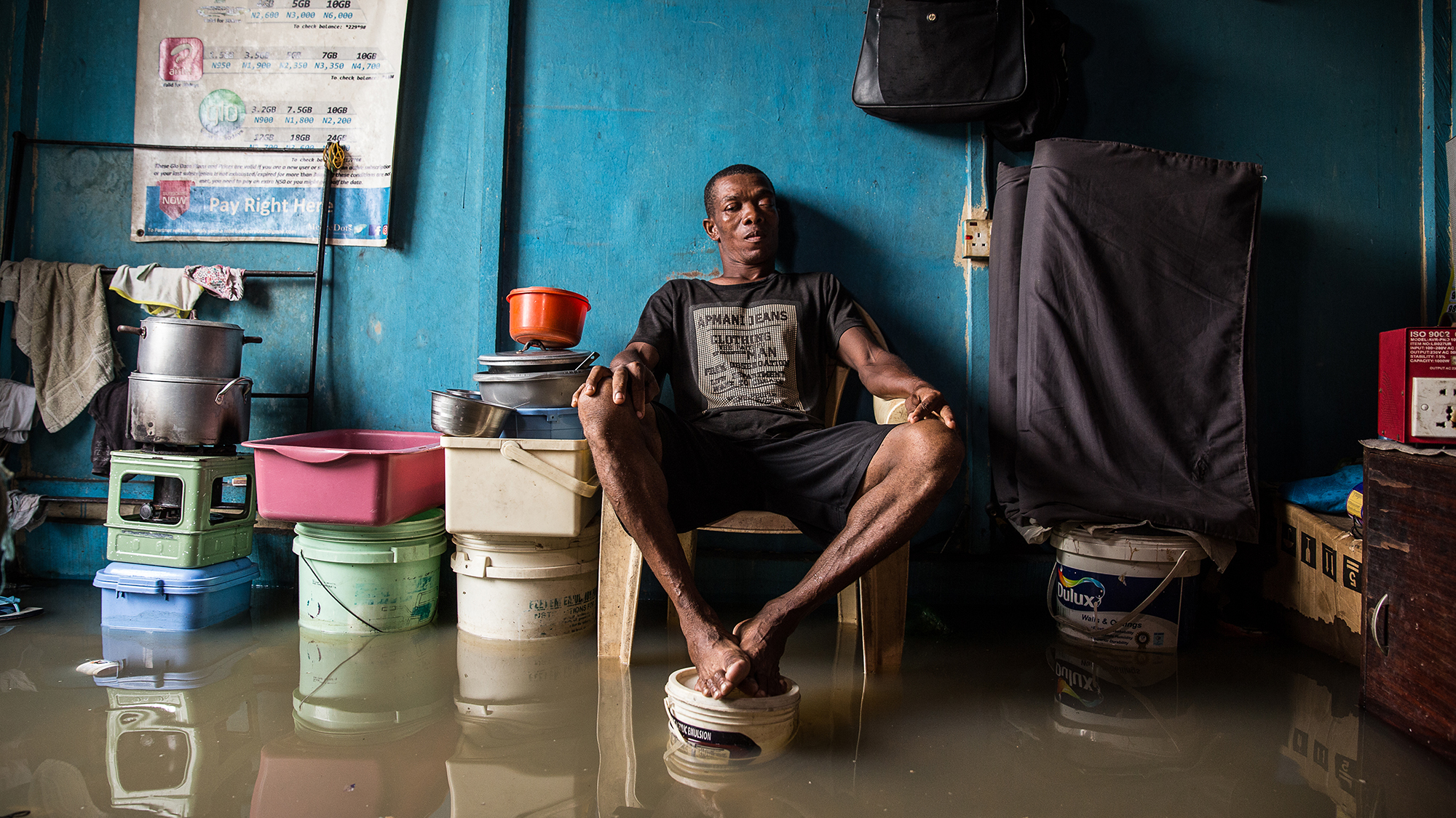 A man sits in centre frame, surrounded by rising flood water and his belongings
