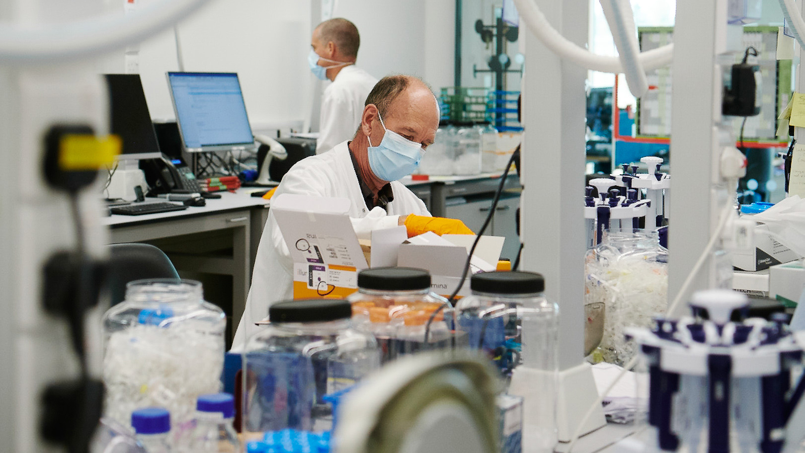 Two scientists wearing facemasks work in a lab, surrounded by lab equipment.