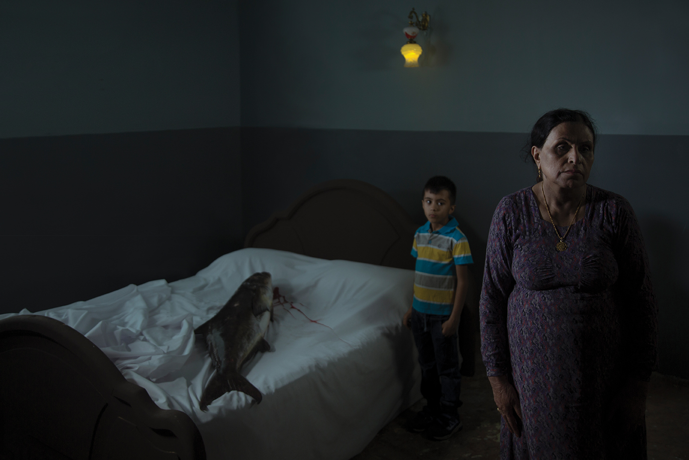 A woman and her son stand in a room, a big fish on the bed.