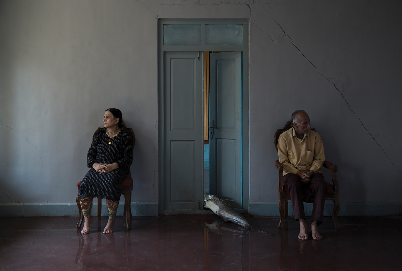 A man and a woman both sit beside an open door, with a big fish on the ground between them. 