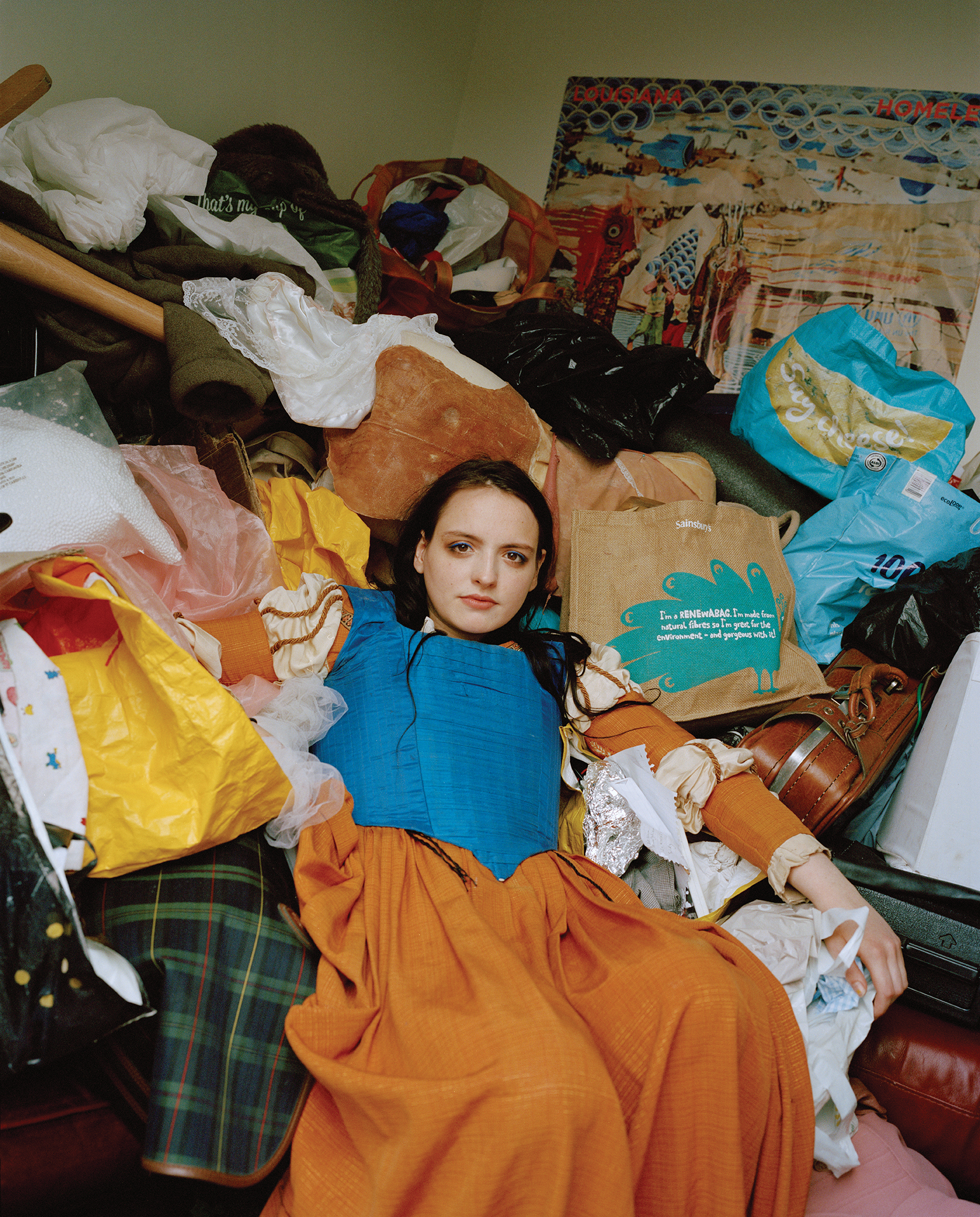 A woman sits on a bed crowded with clothes and bags.
