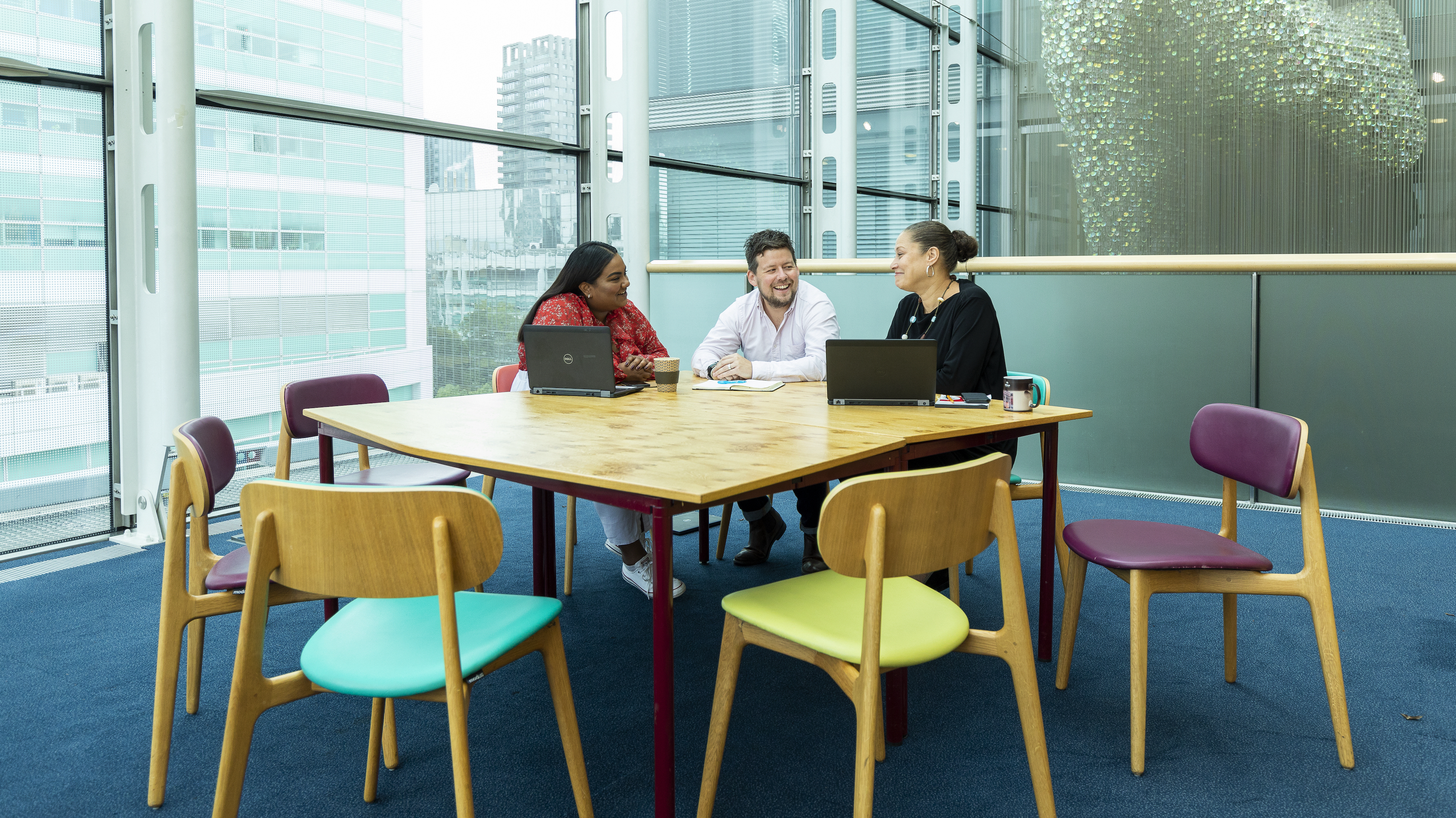 Three Wellcome staff members are sat laughing at a table with laptops. A large window overlooks the city behind them.