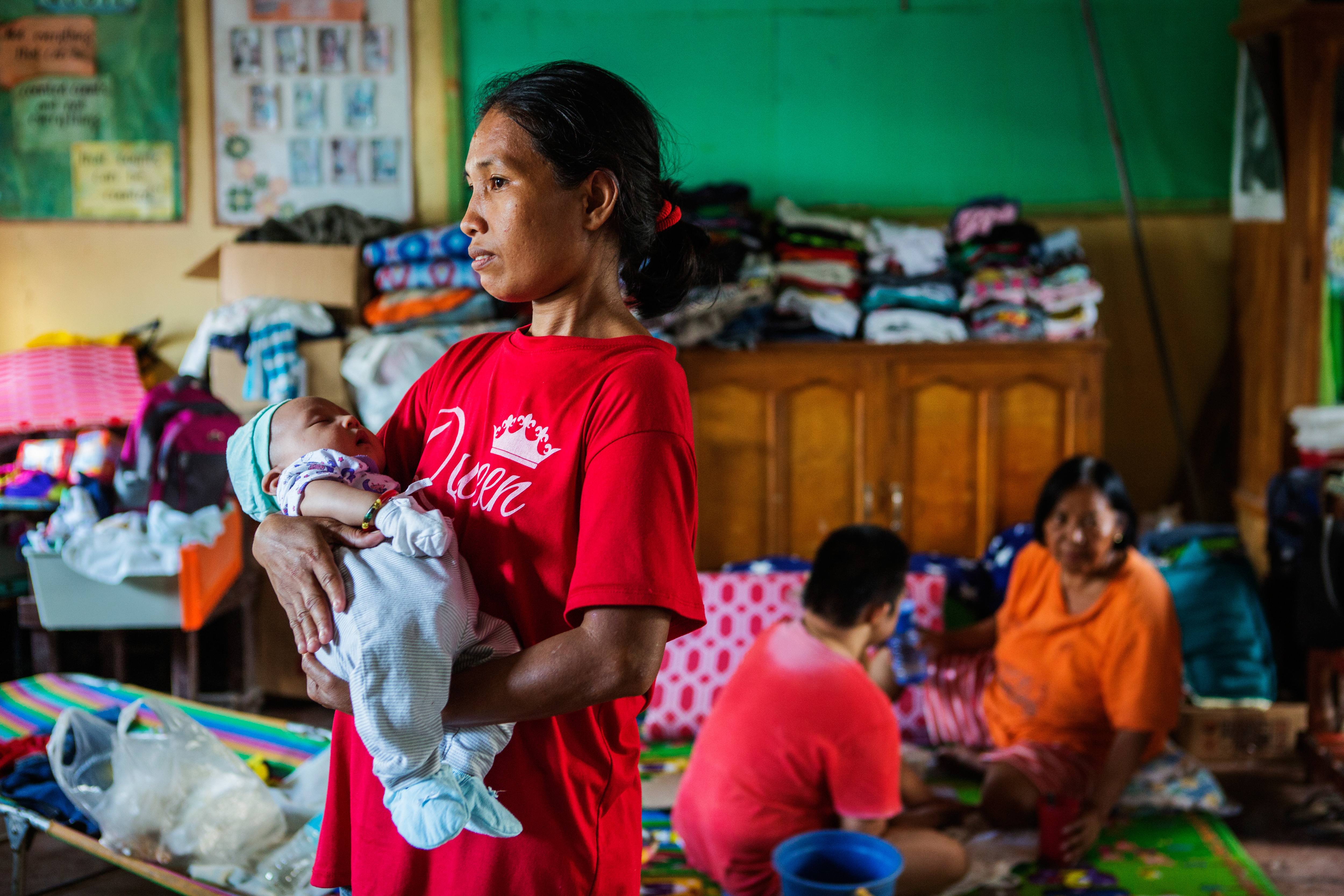 A woman in the Philippines holds her baby standing in a crowded room. Two other people sit behind her, surrounded by belongings.