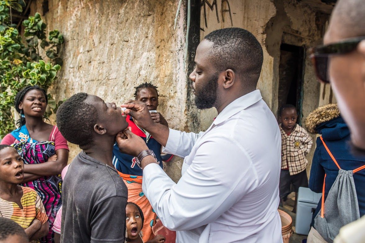 A man in a lab coat is pouring a dose of oral cholera vaccine into the mouth of a man in a black t-shirt in Nampula, Mozambique. They are outside, surrounded by a few women and children who are watching.