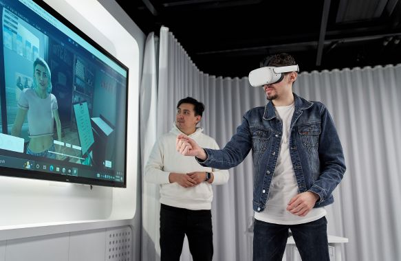 A man wearing a virtual reality (VR) headset standing in front of a TV screen. His hand is outstretched, controlling an animation of a woman on the screen using the VR technology. Another man is standing in the background, watching the screen and the participant's VR control.