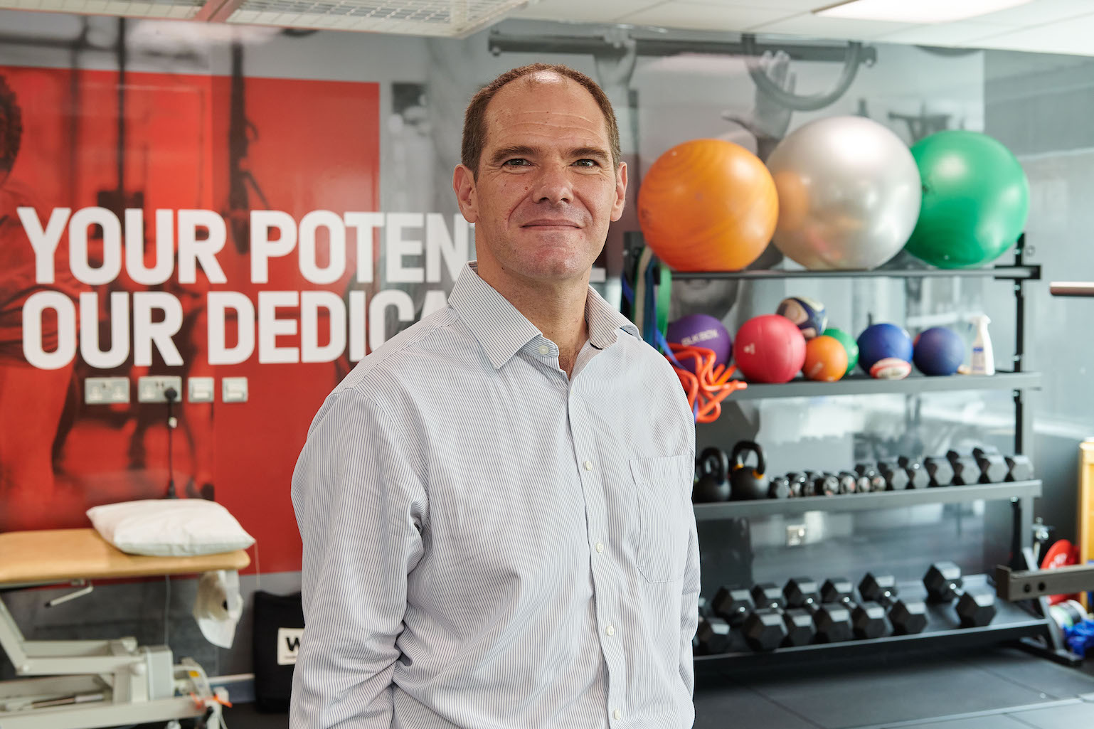 Professor Jonathan Roiser smiles at the camera. He is in a gym, standing in front of a shelf of exercise balls and weights.