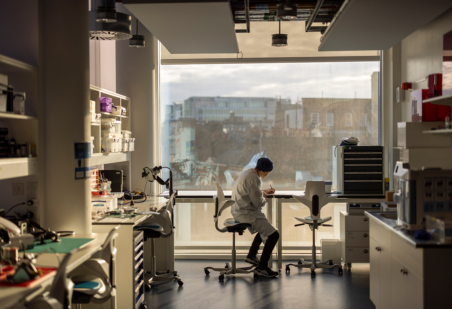 A Laboratory Technician sits in front of a large window, wearing a white lab coat and writing notes in a lab at the Sainsbury Wellcome Centre in London, UK.