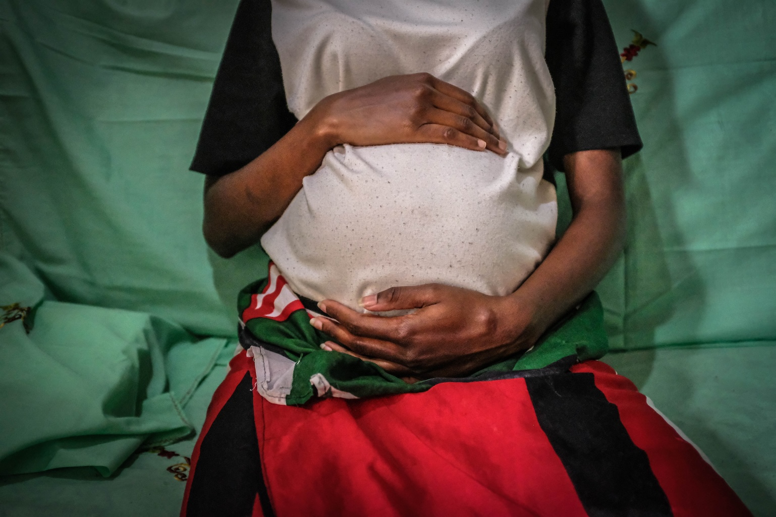 Mercy Kwamboka, 19, is pregnant and holding her baby bump in Nairobi. Mercy is wearing a white t-shirt and a long red skirt.