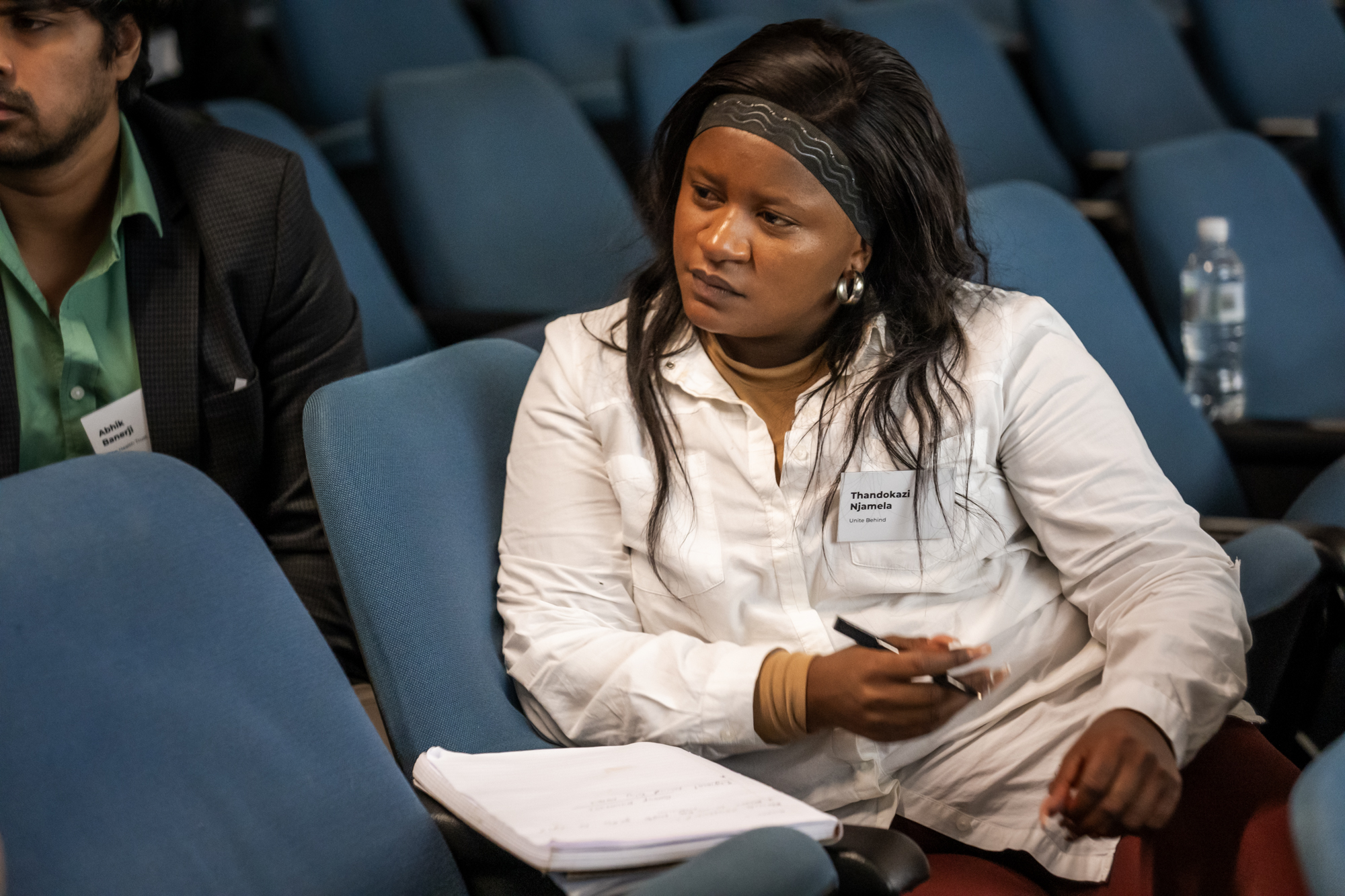 A woman with long hair wearing a white shirt and holding a pen with a notebook in front of her, listening to someone speak.