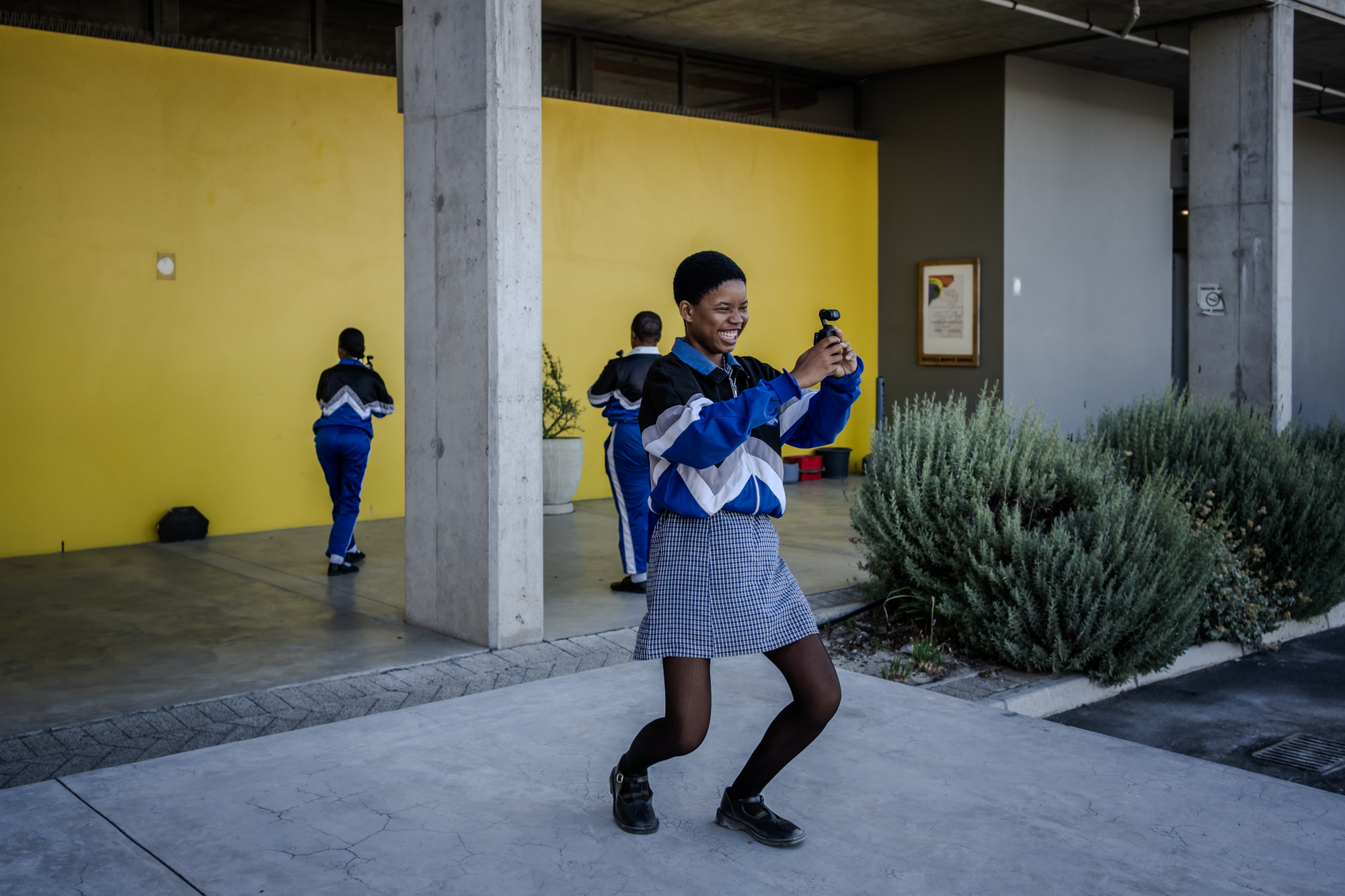 A learner wearing a skirt and long-sleeved jacket, knees bent slightly, is smiling as they take videos with a small camera.