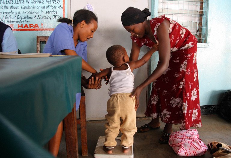 A child has her weight taken at a vaccination ward. She is being helped onto the scale by her mother and a healthcare worker.