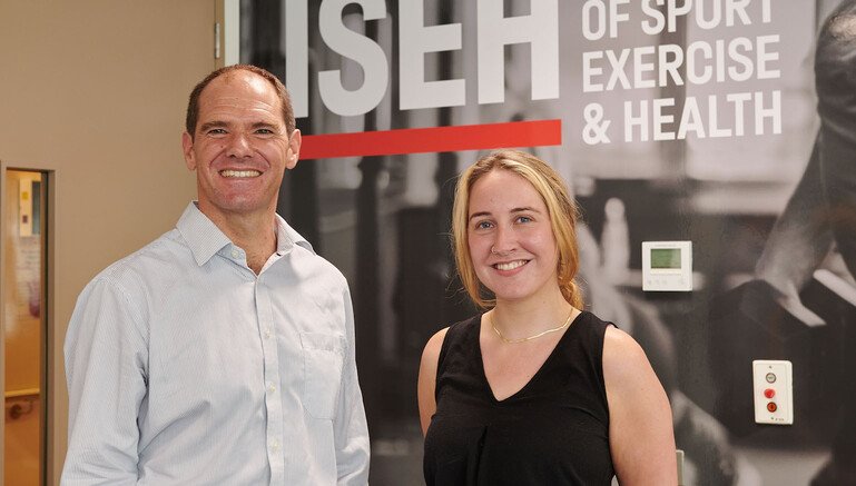 Professor Jonathan Roiser and Emily Hird stand side by side smiling in front of a sign that says 'ISEH - Institute of Sport & Exercise Health'.