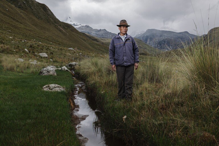 A person stands in a mountainous rural landscape with a small stream to their left.