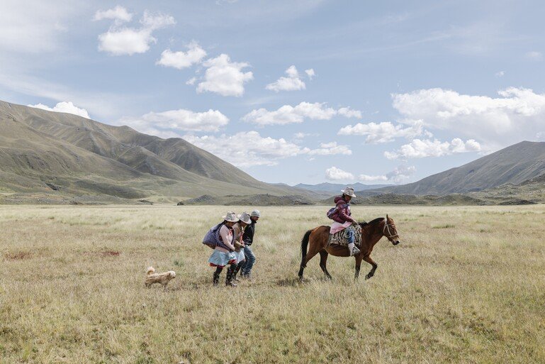 In the dry plains a farmer rides their horse with three others following behind with a small white dog trailing.