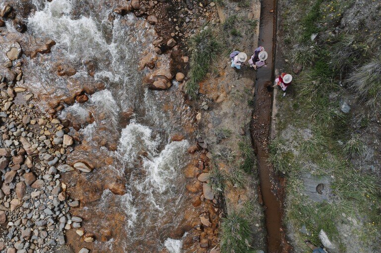 A bird's eye view shows three farmers standing in-between a larger river to their left and a small stream to the right.