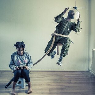 A father dressed as an astronaut floats and is tethered to his daughter who is sitting looking at her phone
