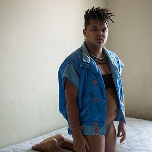 Téo, a transgender man in Brazil, kneels on a bed