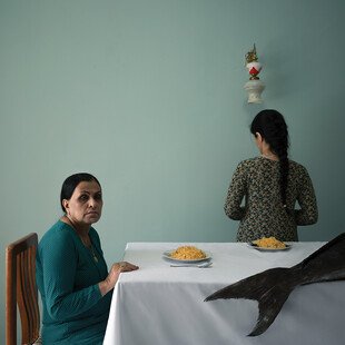 A woman sits at a dinner table, a big fish on the table, while a younger girl stands with her back to the camera.. 