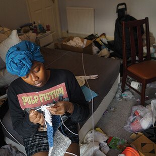 A woman sits on a chair in a cluttered room and knits. 