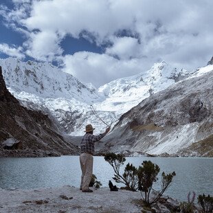 A man stands with his back to the camera in front of a lake. His arm is raised up, holding a rope as he throws something out to the lake. Snow covered mountains surround the lake in the background.