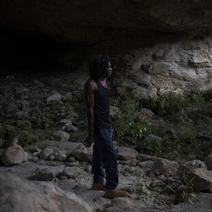 A man stands in a cave surrounded by rocks and some green shrubs. He stands side-on to the camera and looks straight ahead.