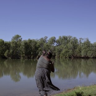 A woman stands next to the river with her back to the camera. She leans forward her arms reaching out as she throws a fishing line into the water. The water is calm and still and trees line the opposite riverbank.