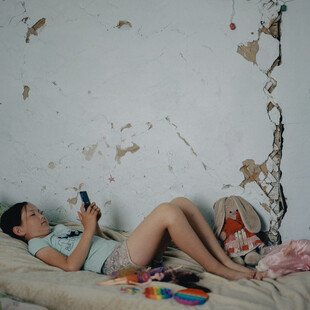 A young girl lies on a bed looking at a mobile phone. Dolls and fidget toys are on the bed next to her. Damaged, cracked walls are visible beside her.