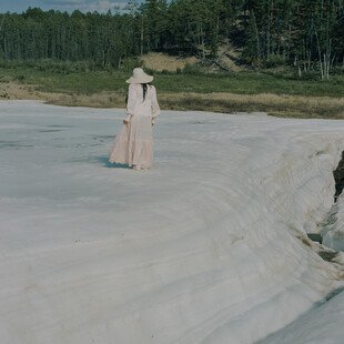 A woman wearing a white hat and long white dress with long sleeves stands on the middle of a large sheet of ice. She stands with her back to the camera and looks out towards green grass and green trees in the background beyond that.