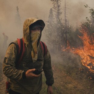 A man stands with his hood up wearing a cloth face covering. He holds a walkie-talkie in one hand and wears a rucksack on his back. Bright orange flames burn trees next to him and smoke fills the air all around. Another person is partially visible behind him in the background.
