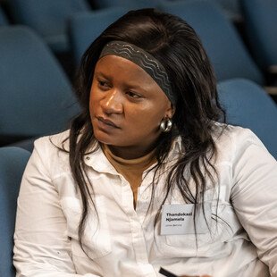 A woman with long hair wearing a white shirt and holding a pen with a notebook in front of her, listening to someone speak.