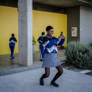 A learner wearing a skirt and long-sleeved jacket, knees bent slightly, is smiling as they take videos with a small camera.