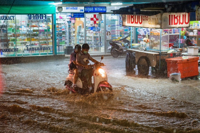 Two people on a motorbike drive through a flooded road as rain falls. Street stalls and shops can be seen in the background.