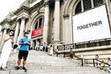 People wearing protective masks stand on the steps of the Metropolitan Museum of Art in New York which displays a banner with the word 'together'.