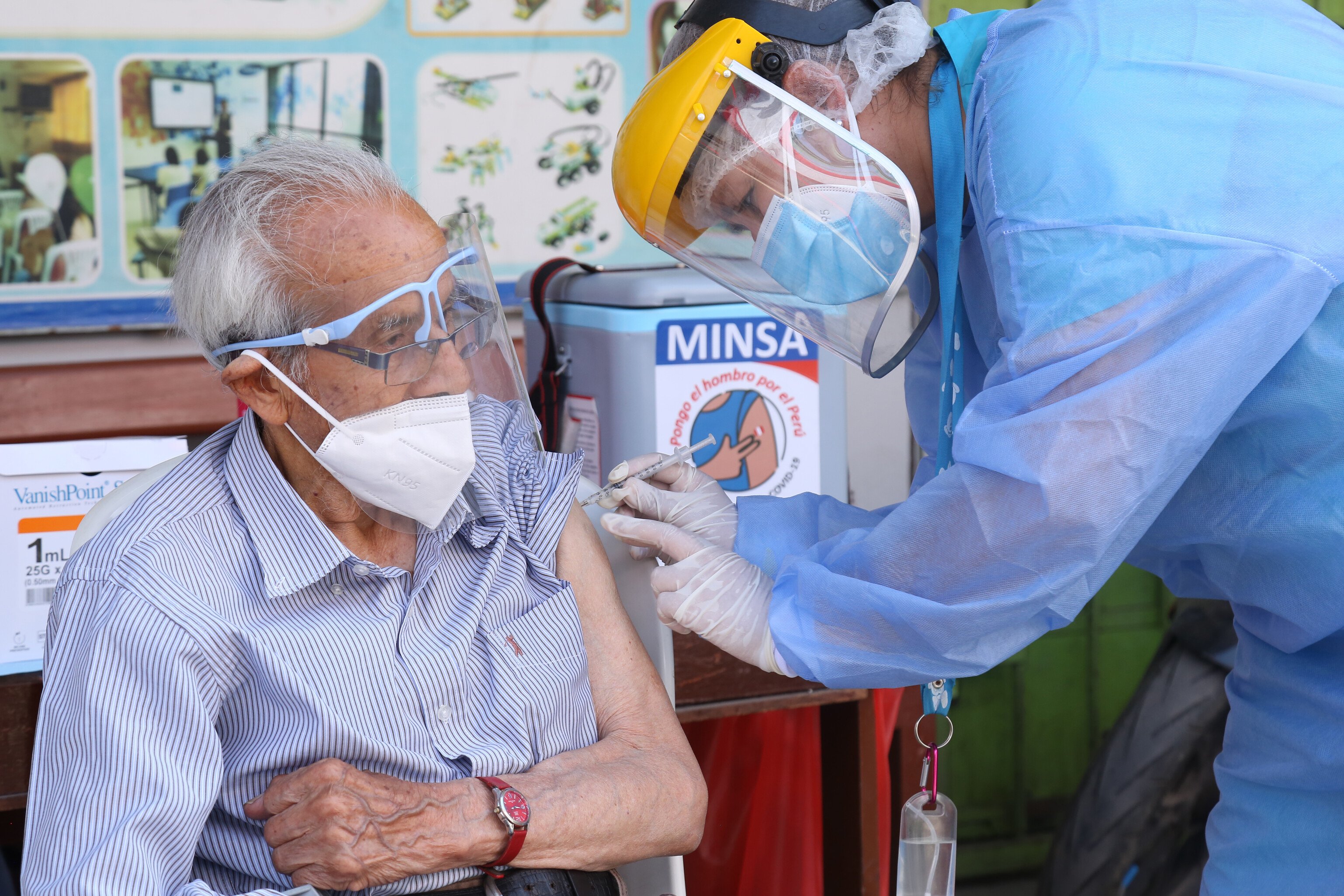 In Peru, a healthcare worker wearing PPE administers a Covid-19 vaccine to an older man wearing a mask and visor.