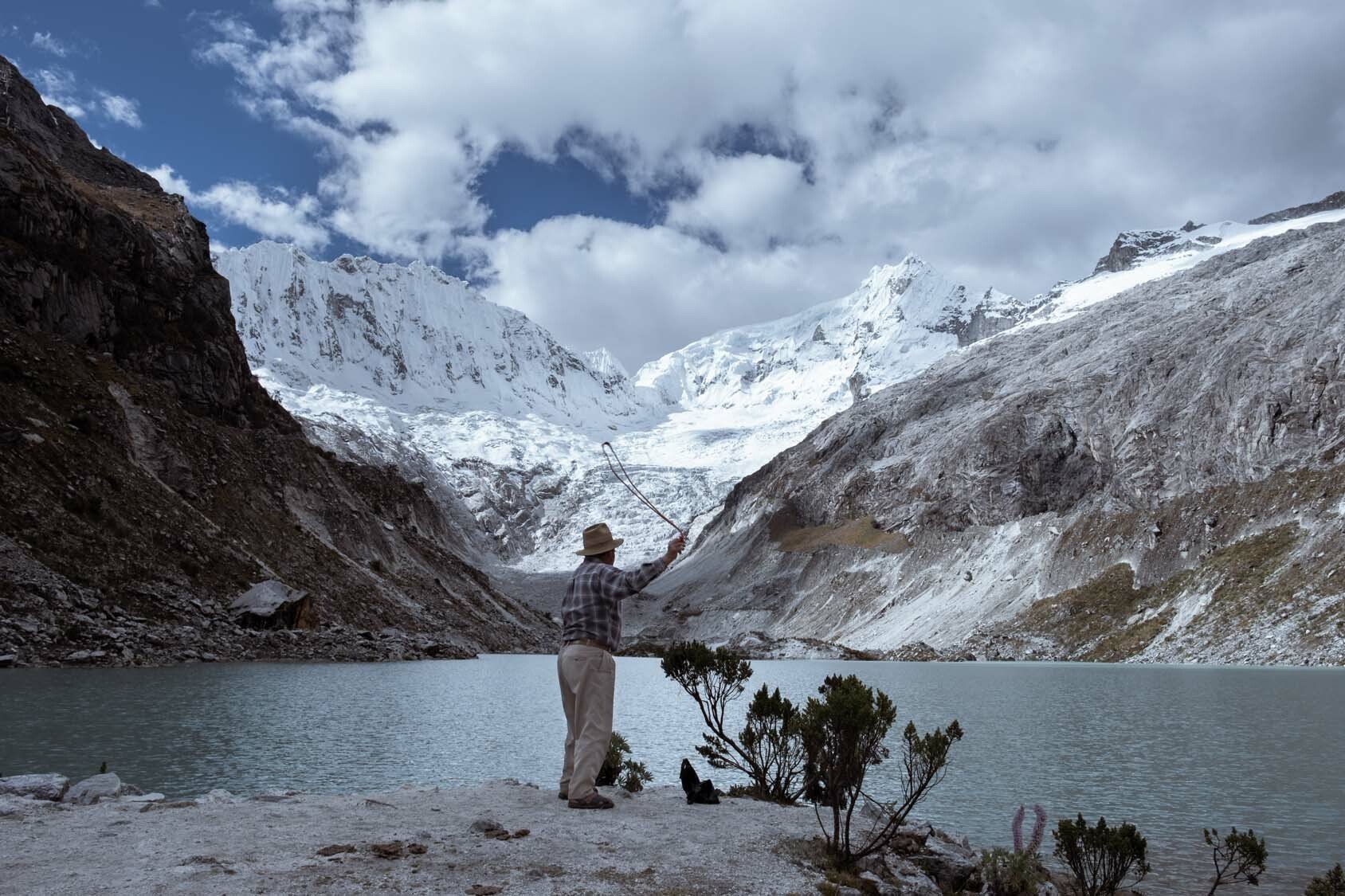 A man stands with back to the camera throwing something out into a lake. The lake is surrounded by snowing mountains.
