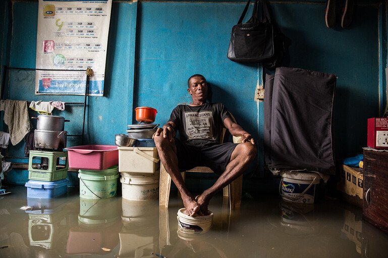 A man sits in his room with his feet up on a paint bucket to avoid the floodwater