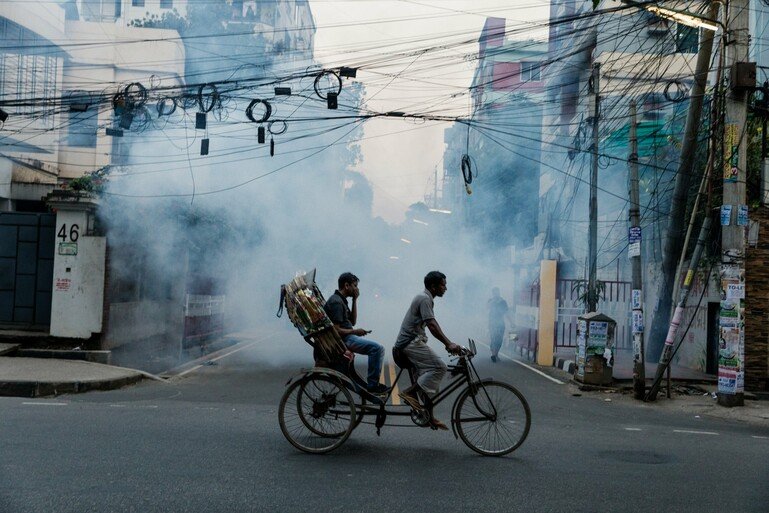 Two people cycling on a street in Uttara which is being sprayed with insecticide to kill mosquitoes