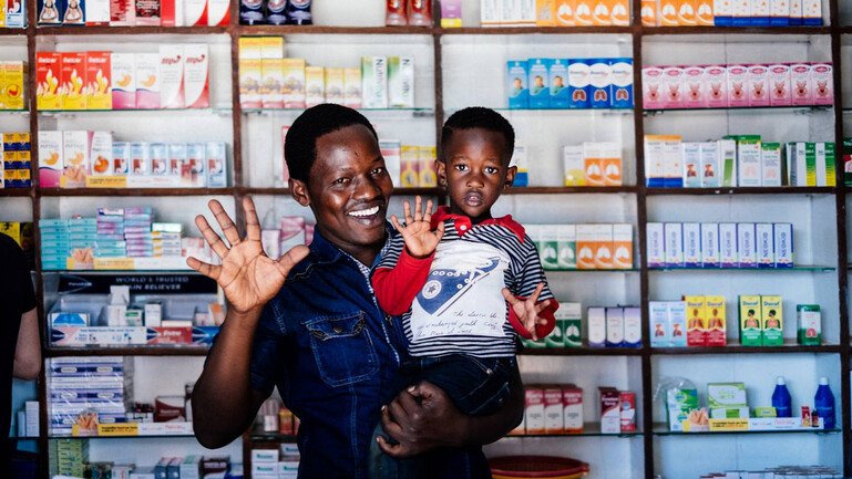 A man stands centre frame in front of a pharmacy. He has a young boy sat on his hip. They are both smiling and waving to the camera.