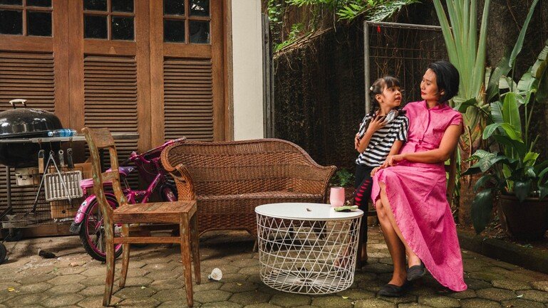 A woman in a long pink dress sits right of frame. A young girl sits on her lap. They look towards one another. 