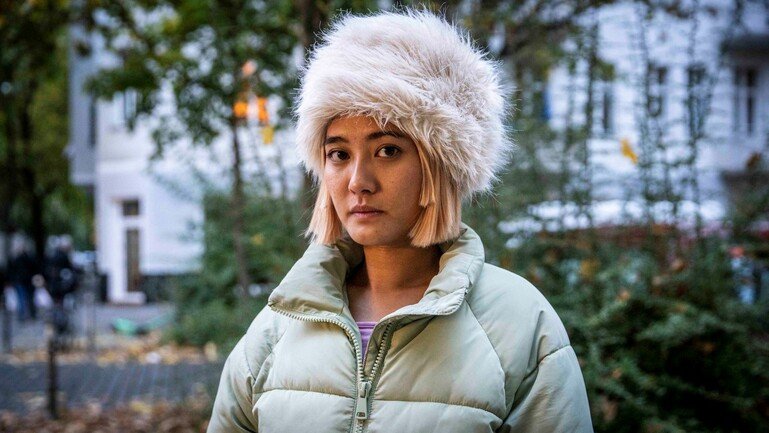 A young woman in a large light green duffel coat stands centre frame. We see her from the chest up. she has a large fur hat on and stares directly into the camera