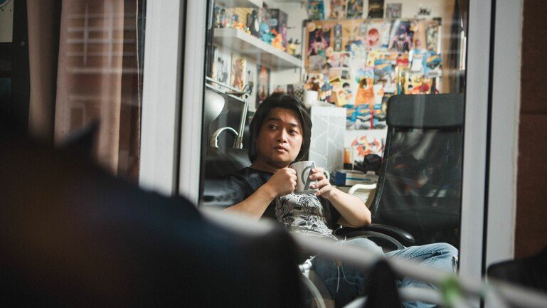 An image of a man sitting down in a room and holding a mug. He is seen from the waist up and looking out of a glass window.