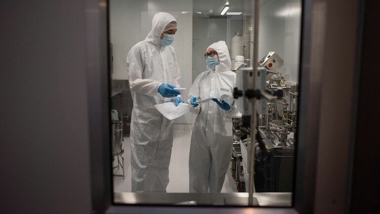 Two scientists in full protective gear stand in a lab discussing lab samples they are holding.