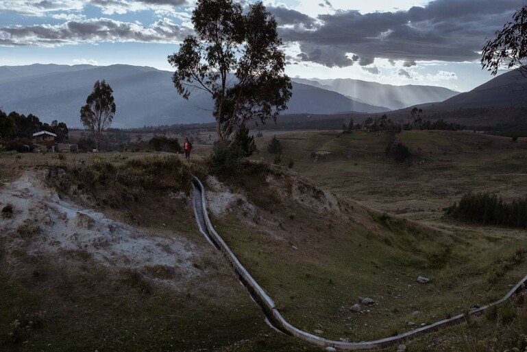 A small canal runs cuts through a landscape. A figure stands at the top of the canal next to a tree.