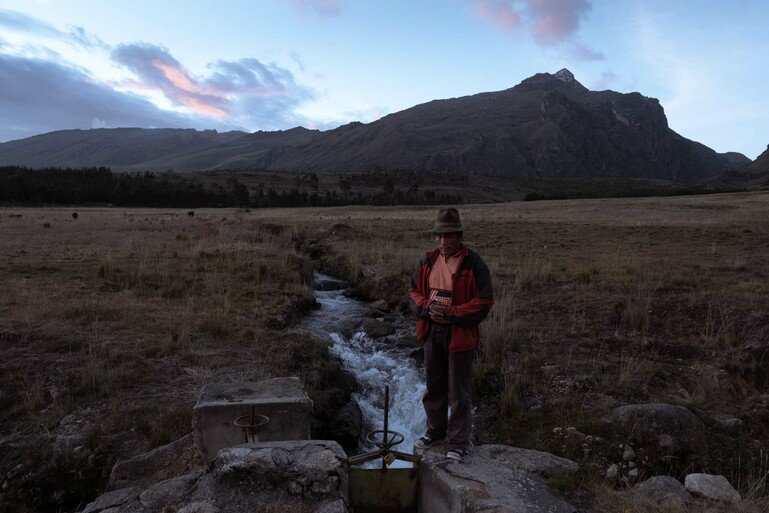 A small river runs through the centre of the frame. A man stands to the right of the river in a plaid red jacket and orange t-shirt.