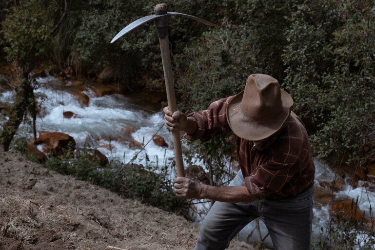 A man is to the right of the frame, he is looking to the grown and has a pic axe aimed down at the ground. A river flows behind him.