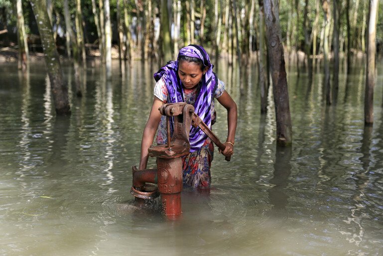 A woman in a purple head scarf stands knee-deep in flood water collecting safe drinking water from a metal hand pump.