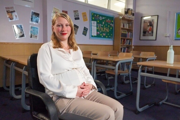 A pregnant woman sits in a classroom looking to camera