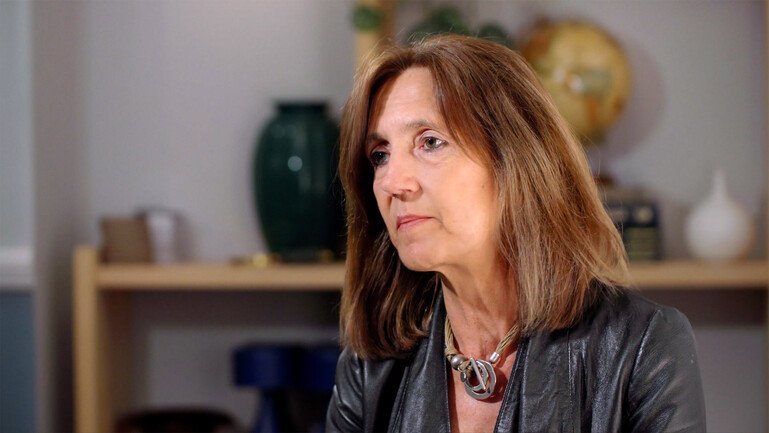 Professor Melissa Leach speaking during an interview, seated indoors with shelving and a globe visible in the background.