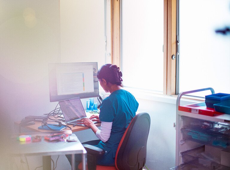 A healthcare professional in blue clinical scrubs sits at a desk using a laptop and desktop computer in a hospital office.